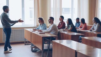 Male professor in casual clothes is talking to group of students sitting at tables in classroom and making notes. Large lecture hall with desks, chairs and large windows is visible. - obrazy, fototapety, plakaty