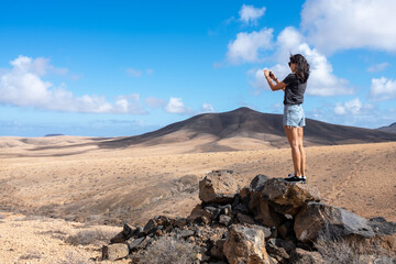 Young female traveler and smartphone photographing beautiful mountains on sunny day on Fuerteventura island, Spain
