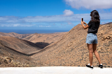 Young woman traveler with smartphone photographing the landscape of the island of Fuerteventura on a sunny day with the ocean in the background, Spain