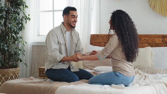Hispanic Indian man and woman sitting on bed in new home celebrate buying real estate relocation. Boyfriend and girlfriend husband and wife couple in bedroom talking laughing talk funny holding hands