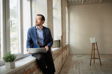 Portrait of young businessman sitting at the window with laptop and working on new startup. Career...