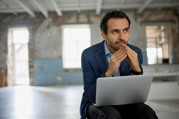 Portrait of man, businessman, worker sitting with laptop with thoughtful expression in big empty room. Professional brainstorming