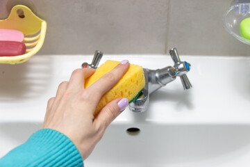 Woman's hand washes the faucet in the sink with a yellow sponge, cleaning the faucet in the...