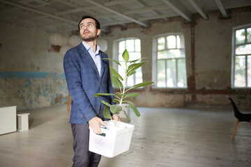 Portrait of young businessman walking out empty room with flower. Business closure. Depression and crisis