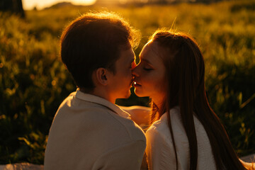 Close-up silhouette view of loving young couple sitting hugging and kissing on beautiful meadow with green grass having picnic date on background of warm sunlight, in summer evening sunny sunset.