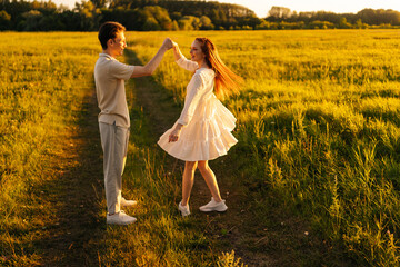 Beautiful young woman in dress dancing with loving boyfriend on green meadow in summer evening...