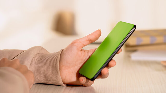 Partial view of woman holding cellphone with green screen at home.