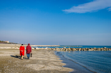 Due persone vestite di rosso camminano lungo la spiaggia del Lido di Venezia in una giornata invernale di sole