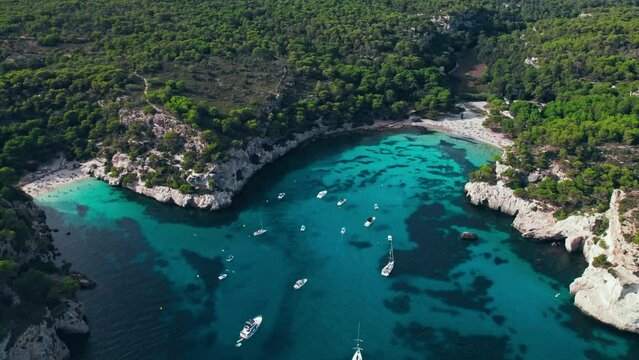 Aerial drone view of people having a good time by the beach with turquoise water. Tourists enjoy Cala Macarelleta and Macarella on the Balearic Islands, Spain, swimming and sailing yachts and boats.