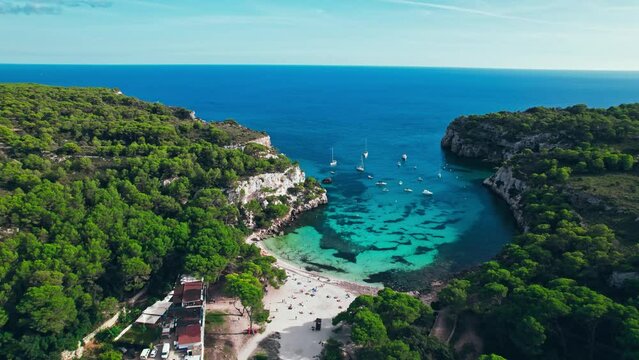 Aerial drone view of people having a good time by the beach with turquoise water. Tourists enjoy Cala Macarelleta and Macarella on the Balearic Islands, Spain, swimming and sailing yachts and boats.