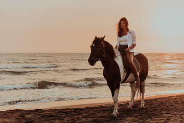 Fototapeta na wymiar Woman in summer clothes enjoys riding a horse on a beautiful sandy beach at sunset. Selective focus 