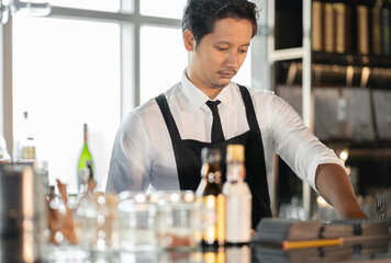 Asian bartender man standing at bar counter prepare to service cocktail alcohol at restaurant, portrait. Handsome young barman in uniform working at nightclub. Waiter person in nightlife lifestyle.