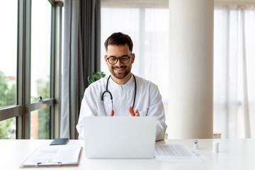 Portrait of man doctor talking to online patient on laptop screen sitting at clinic office desk...