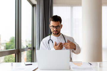 Concentrated young physician working on computer looking at laptop screen thinking on problem solution concept. Serious man doctor wearing white coat doing online research at workplace.