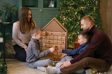 Advent calendar in the form of a house with thirty-one boxes with gifts for every day of December. Family opens presents with sons at Christmas tree