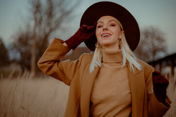 Happy smiling fashionable woman wearing trendy autumn outfit with classic brown coat, hat, suede gloves, beige cashmere turtleneck sweater, hoop earrings, posing outdoor. Copy, empty space for text
