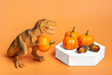 Toy dinosaur Tyrannosaurus holding pumpkin in its paws, on white podium isolated on orange...