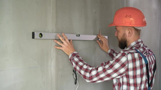 male engineer or architect in a protective helmet measures the level of the wall. A builder in a house is checking a flat concrete wall. Repair and construction