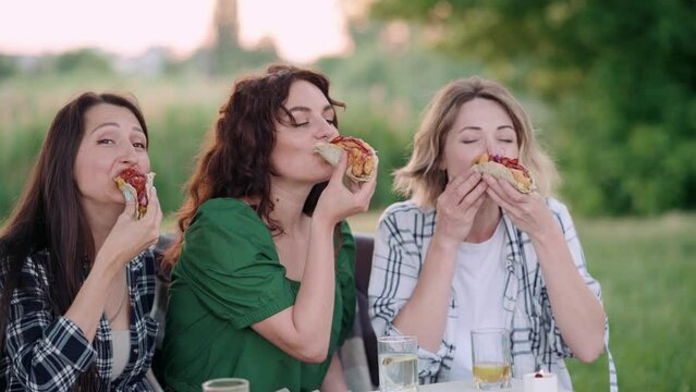 Three young Caucasian women eat tacos in nature with great appetite.