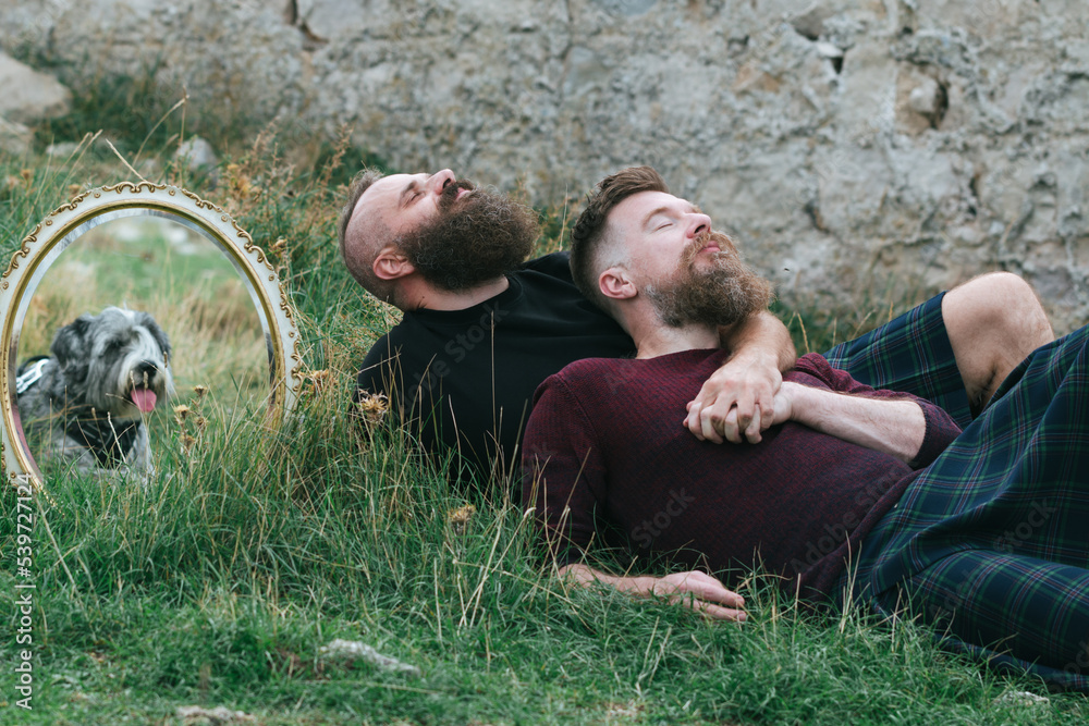 Sticker Hipster fashionable  gay couple wearing kilts having fun in grass.  Long beard male homosexual partners lying together outside, holding hands, enjoying in romance outdoors with their dog.