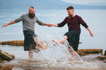 Attractive gay couple in kilts splashing water and having fun on the beach. Homosexual long beard...
