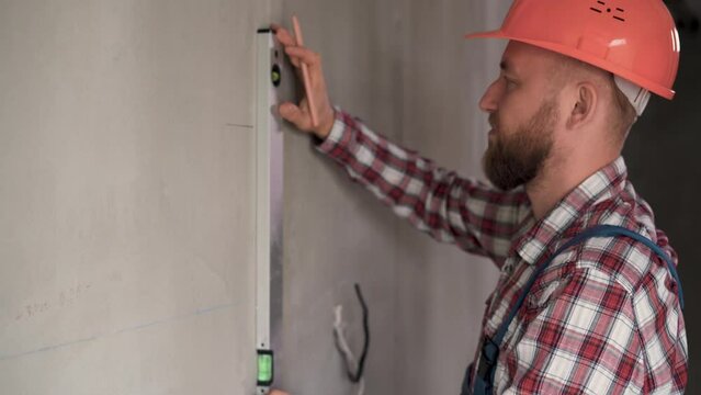 male engineer or architect in a protective helmet measures the level of the wall. A builder in a house is checking a flat concrete wall. Repair and construction