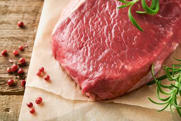 Raw beef steak. Fresh beef rib eye steak with fork rosemary, salt and pepper on piece of parchment paper on old wooden background. Top view. Mockup for design idea.