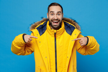 Young surprised shocked amazed fun cool caucasian man 20s in yellow down jacket point index fingers on himself isolated on plain blue color background studio portrait. People winter lifestyle concept.