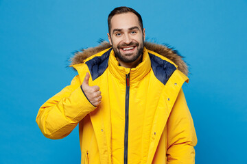 Young smiling fun happy satisfied cheerful man 20s wearing yellow down jacket showing thumb up like gesture isolated on plain blue color background studio portrait. People winter lifestyle concept