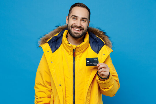 Young smiling cheerful fun cool rich caucasian man 20s wearing yellow down jacket hold in hand credit bank card isolated on plain blue color background studio portrait People winter lifestyle concept