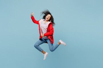 Full body fun young woman of African American ethnicity 20s she wear red jacket jump high do playing guitar gesture isolated on plain pastel light blue cyan background Wet fall weather season concept