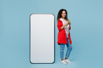 Full body young woman of African American ethnicity in red jacket near big huge blank screen mobile cell phone workspace copy space use smartphone isolated on plain pastel light blue cyan background.