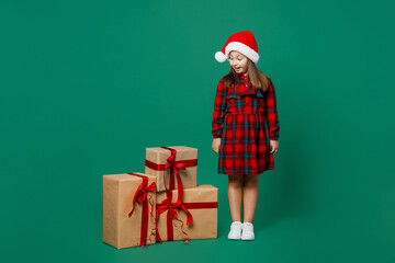 Full size merry little child kid girl 7 year old wear red dress Christmas hat posing look at near present boxes with gift ribbon bow isolated on plain dark green background Happy New Year 2023 concept