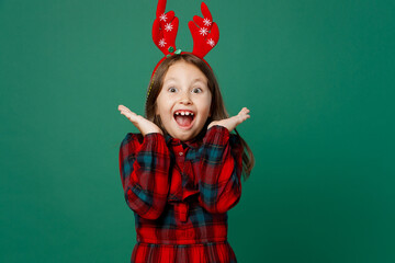 Merry happy little child kid girl 6-7 year old wear red dress Christmas hat posing fun decorative deer horns on head spread hands isolated on plain dark green background. Happy New Year 2023 concept.