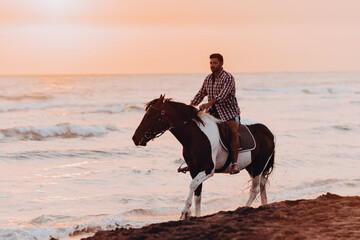 A modern man in summer clothes enjoys riding a horse on a beautiful sandy beach at sunset. Selective focus 