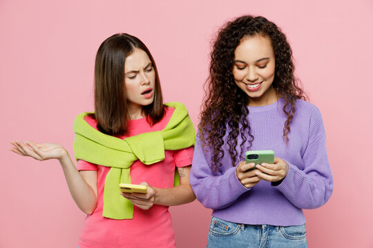 Young two friends shocked confused wondered questioned women wearing green purple shirts together hold in hand use mobile cell phone spread hands isolated on pastel plain light pink color background.