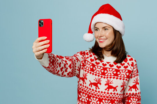 Young fun merry woman 20s wear warm red knitted sweater Santa hat posing doing selfie shot on mobile cell phone isolated on plain pastel light blue cyan background Happy New Year 2023 holiday concept