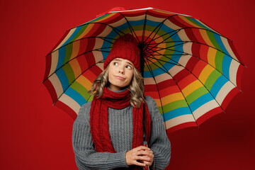 Young sad woman wear grey sweater scarf hat hold opened colorful umbrella look aside on area isolated on plain red background studio Healthy lifestyle ill sick disease treatment cold season concept.