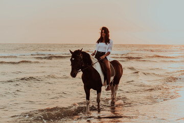 Woman in summer clothes enjoys riding a horse on a beautiful sandy beach at sunset. Selective focus 