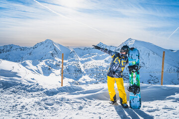 Portrait of a snowboarder with a snowboard showing with hand distant piste on the top of the snowy Pyrenees Mountains. El Tarter, Grandvalira, Andorra - obrazy, fototapety, plakaty
