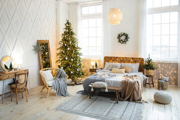 Cozy eco-style bedroom decorated for Christmas. Double bed with pillows and blankets near a large Christmas tree decorated with bright garlands and minimalist white toys in a bright spacious interior