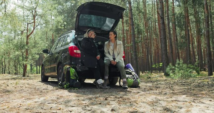 Young satisfied daughter with middle-aged mother sitting in open trunk of car resting drinking tes from thermos talking spending time together family concept cheerful women resting in forest.