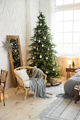 Cozy eco-style interior decorated for Christmas. A rattan chair with a pillow and a plaid near a large Christmas tree with bright garlands and white toys in a white spacious bedroom. Festive interior