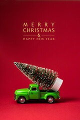 Green retro toy car carrying a Christmas tree on red background with text - Merry Christmas and Happy New Year. Christmas and New Year celebration concept. Greeting card. Copy space, selective focus