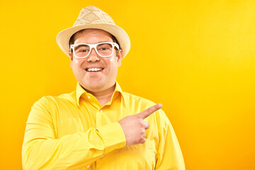 Funny fat man tourist in hat and glasses with surprised face smiling pointing with finger at empty copy space for text or product isolated on yellow background, advertising banner