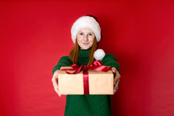 Merry Christmas Portrait of a beautiful young teenage girl in a cozy knitted green sweater and Santa's hat holding gift boxes. The red background is the place for the text.