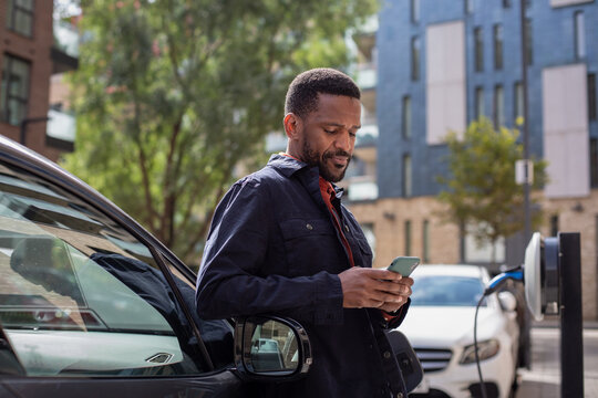 African American Male Charging Electric Car And Using Smartphone App In City