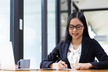 Attractive young businesswoman secretary in office working on laptop writing work schedule on notepad.