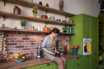 Obraz premium Happy and smiling large woman working on her laptop while sitting over the kitchen table in the large kitchen she looking at the window and smile large