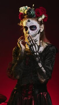 Vertical video: Young scary woman doing hush gesture to keep secret, wearing santa muerte costume and showing slience mute and secrecy sign with finger over lips. Holy dios de los muertos holiday.